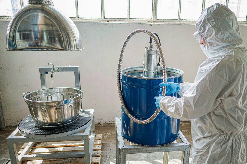 worker in protective coverall decanting clear amine from a blue steel drum under an exhaust hood